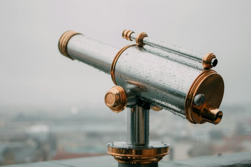 A detailed close-up of a metallic telescope covered in water droplets, set outdoors on a cloudy day.