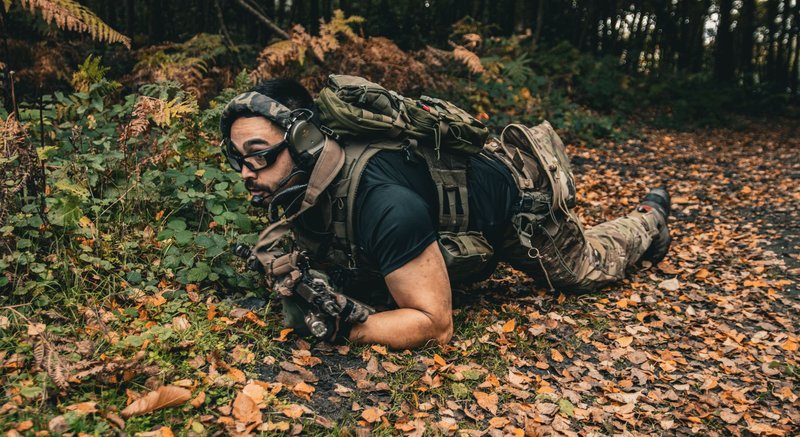 A soldier in full tactical gear crawling through a forest floor covered with autumn leaves.