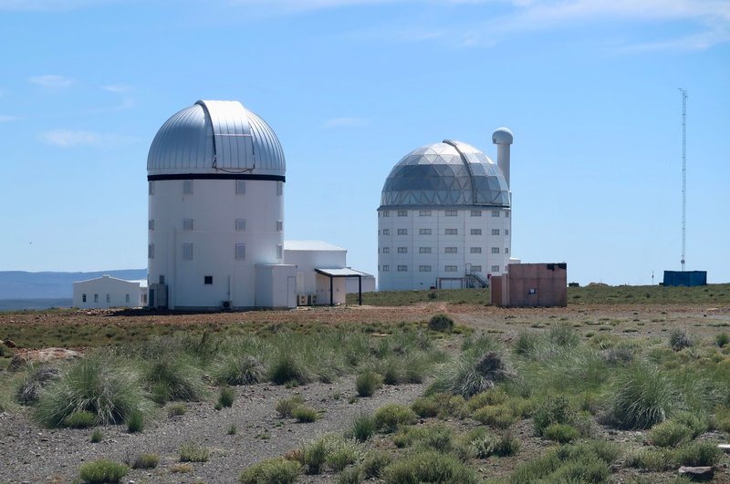 Two observatory domes stand in a sparse, desert-like landscape under a clear blue sky.