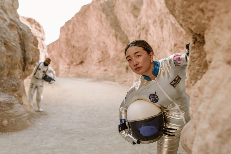 Two astronauts in silver spacesuits explore a canyon-like rocky terrain, evoking a futuristic or Mars landscape.