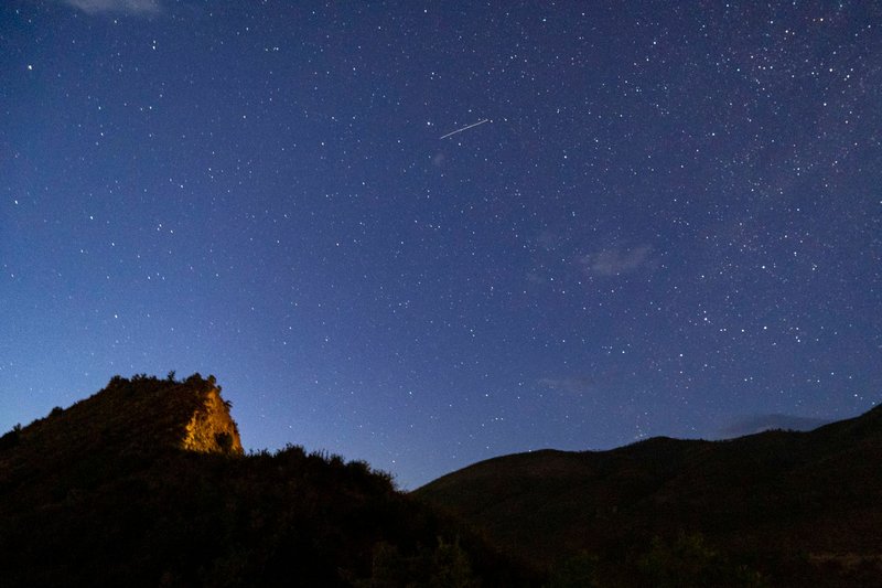 A breathtaking view of a clear starry sky with a meteor streak above a silhouetted mountain landscape at night.