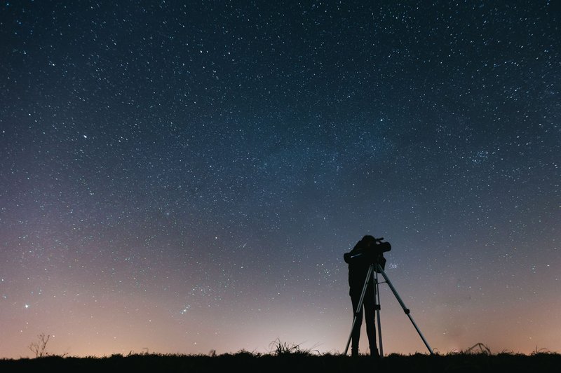 A silhouetted person using a telescope to observe the night sky filled with stars.