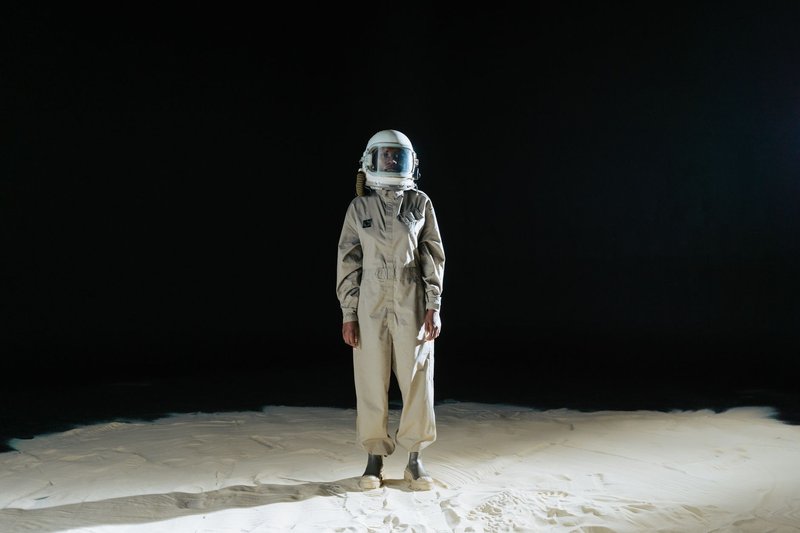 An astronaut standing alone in a space suit on a desert-like terrain against a dark background.